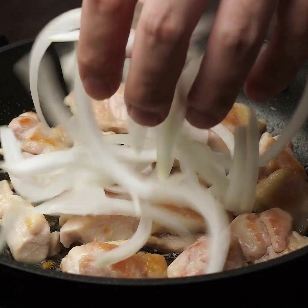 Una mano agregar cebollas blancas finamente picadas en una sartén sobre trozos de pollo parcialmente cocidos.