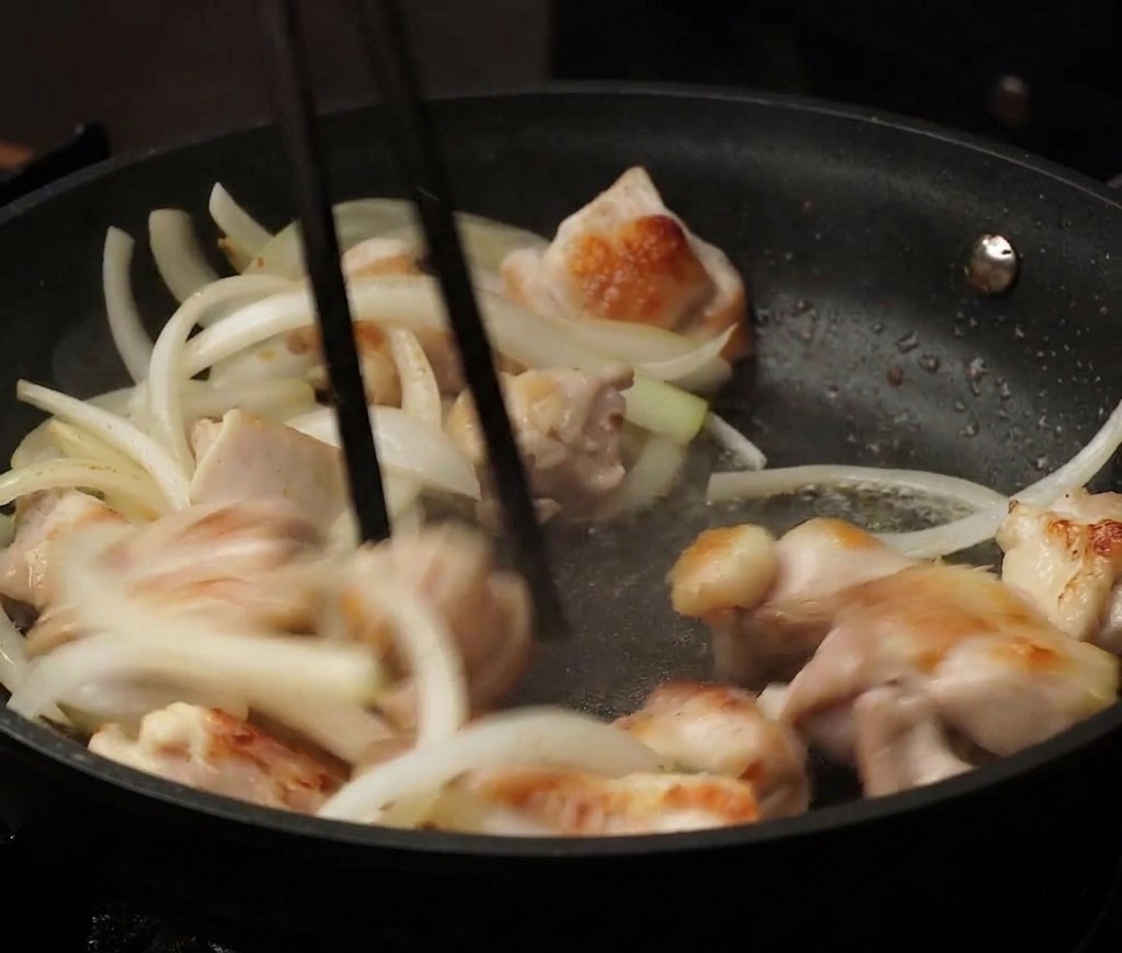 Trozos de pollo del tamaño de un bocado y cebollas en tiras siendo salteados juntos en una sartén antiadherente negra usando palillos.