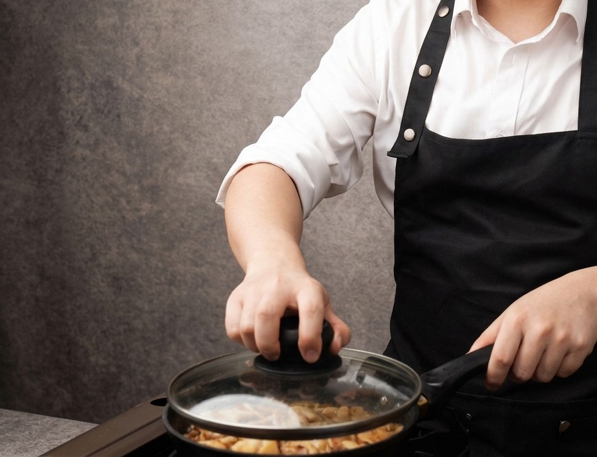 Una mano colocando una tapa de vidrio transparente en una sartén llena de trozos de pollo y tiras de cebolla cocinándose en una salsa de color marrón claro.