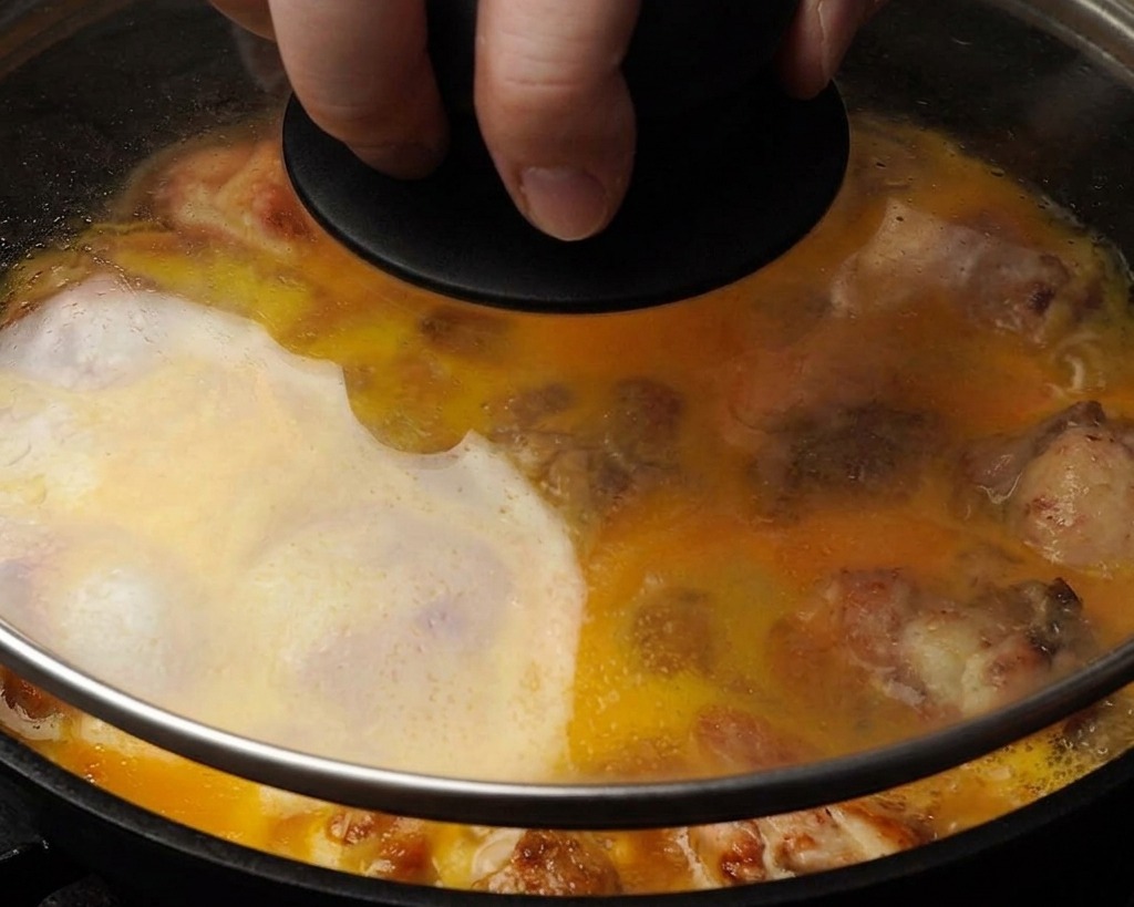 Un plano de cerca de una sartén con tapa de vidrio que muestra el huevo líquido amarillo cubriendo parcialmente los trozos de pollo mientras se cocinan al vapor en el interior.