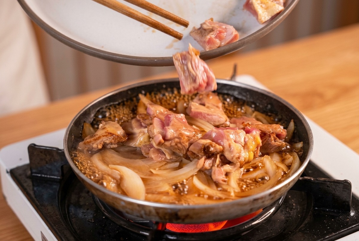 Trozos de pollo sellado siendo añadidos con palillos a una sartén con cebollas hirviendo y caldo dashi.