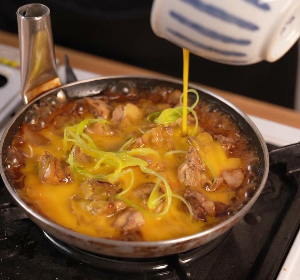 Huevo batido vertiéndose desde un tazón a rayas azules y blancas sobre pollo y cebollín cocinándose a fuego lento en una salsa oscura.