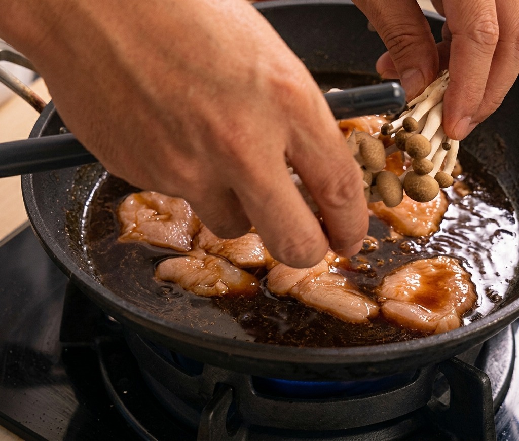 Unas manos colocando un manojo de hongos shimeji en una sartén con pollo hirviendo en salsa oscura.
