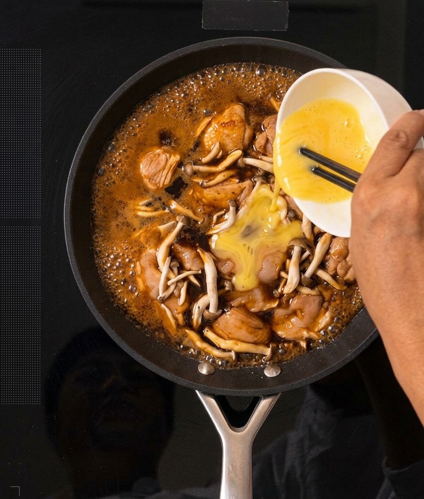 Huevos batidos de color amarillo vertiéndose desde un tazón blanco en una sartén con la mezcla burbujeante de Oyakodon.