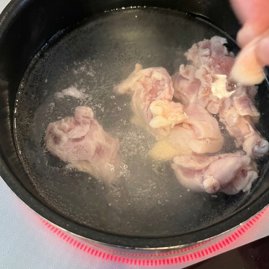 Unas manos introduciendo trozos de pollo crudo en una olla con agua hirviendo sobre una placa de inducción roja.