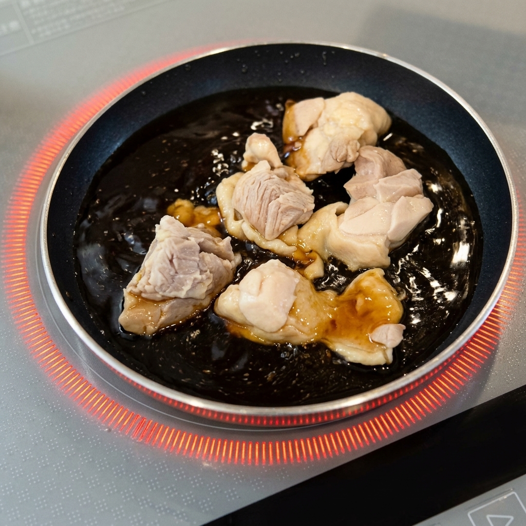 Trozos de pollo escaldados cociéndose a fuego lento en una salsa marrón oscura burbujeante dentro de una pequeña sartén redonda.