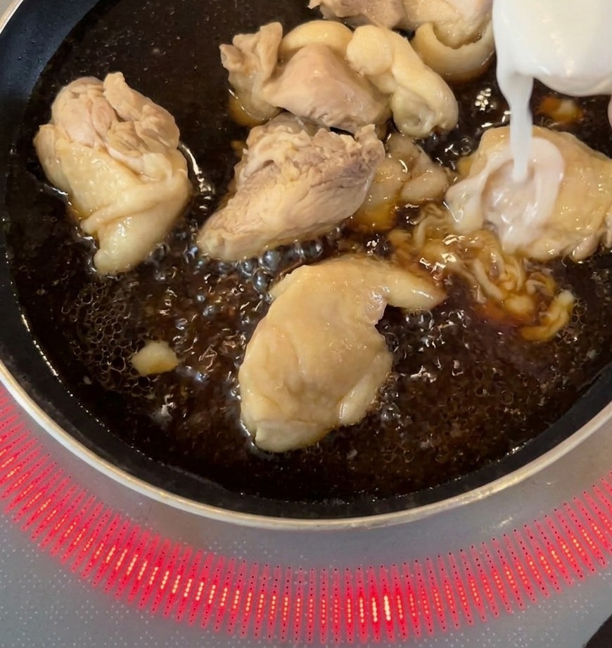 Cebollas blancas laminadas siendo añadidas a una sartén con trozos de pollo cocinándose en una salsa oscura.