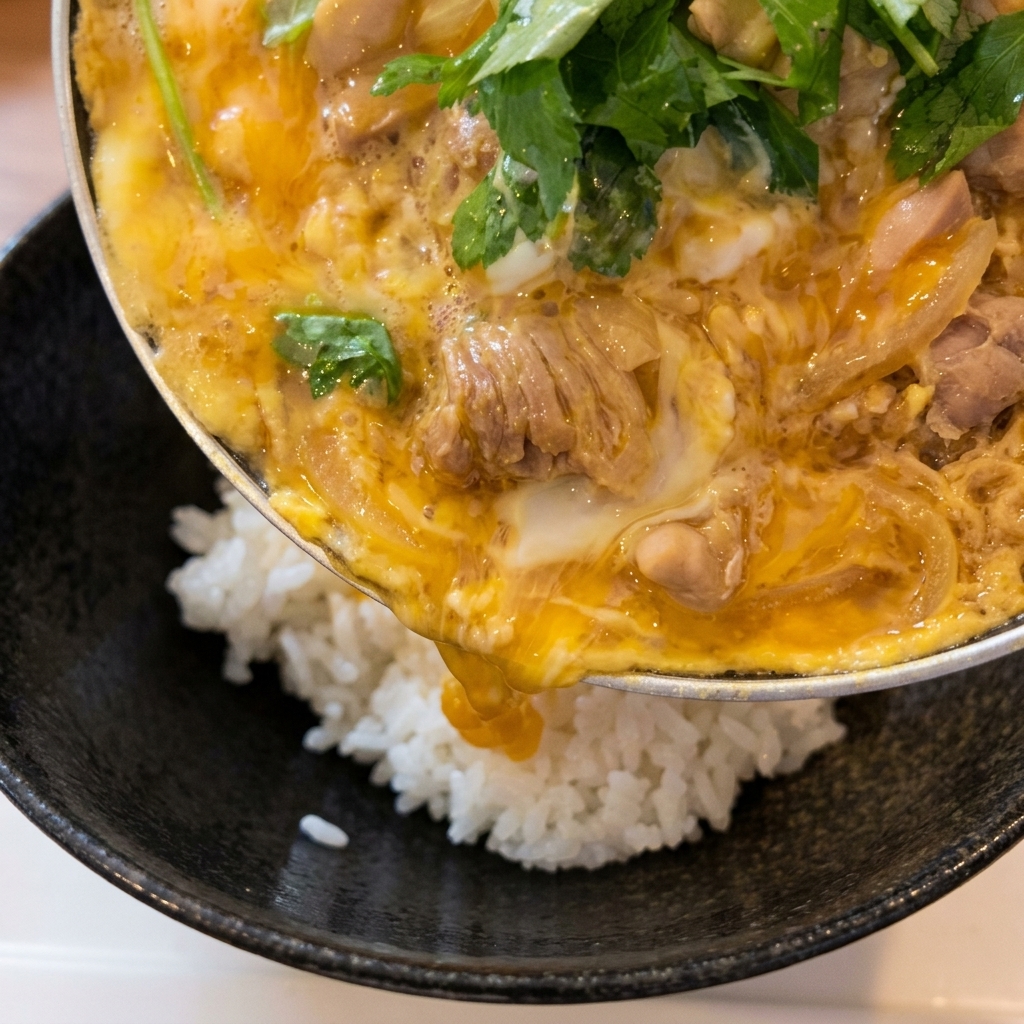 Una sartén inclinándose para deslizar la cobertura de pollo y huevo cocidos sobre un cuenco de arroz blanco esponjoso.