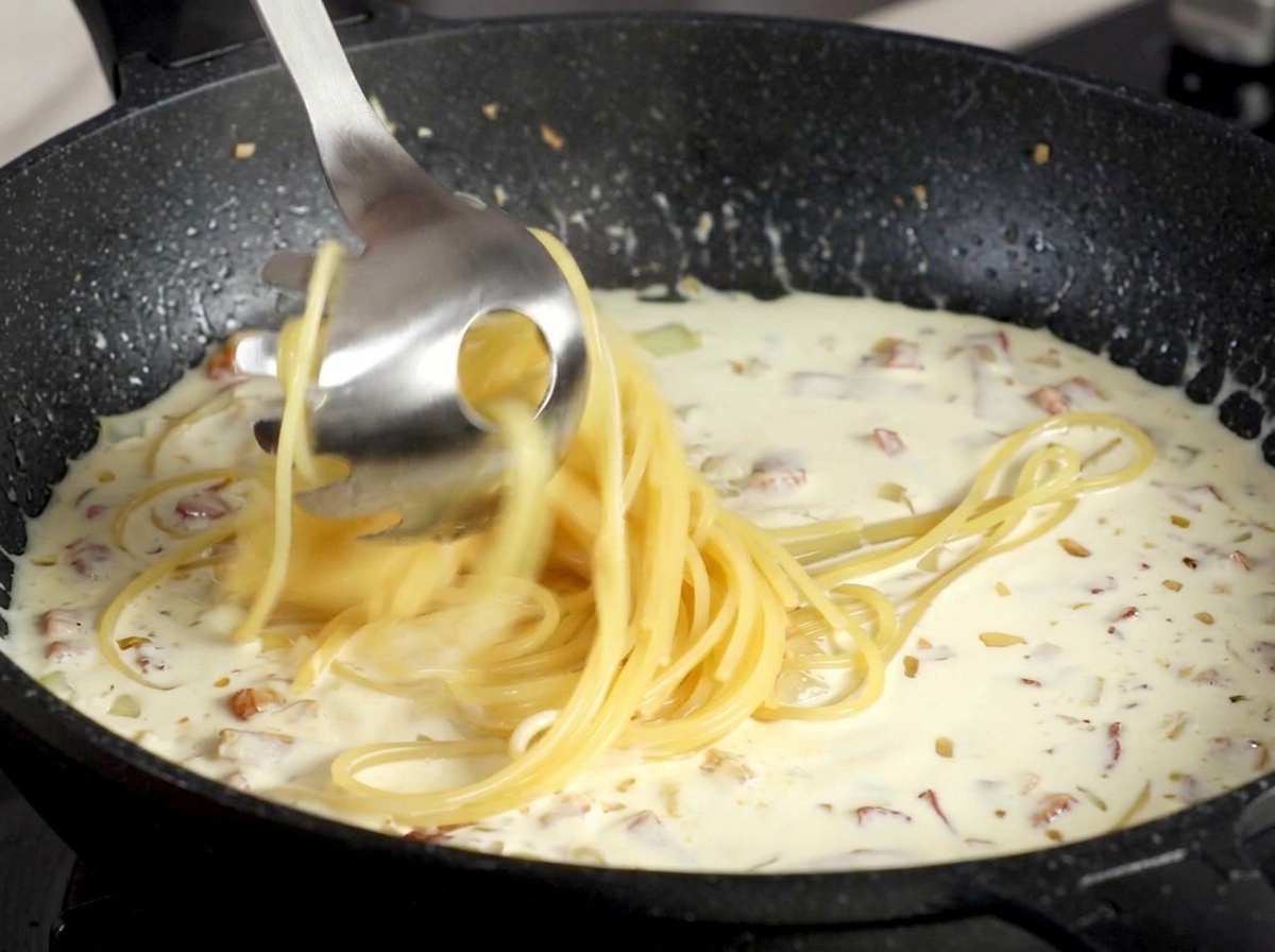 Espaguetis siendo trasladados con una cuchara para pasta a una salsa cremosa de bacon.