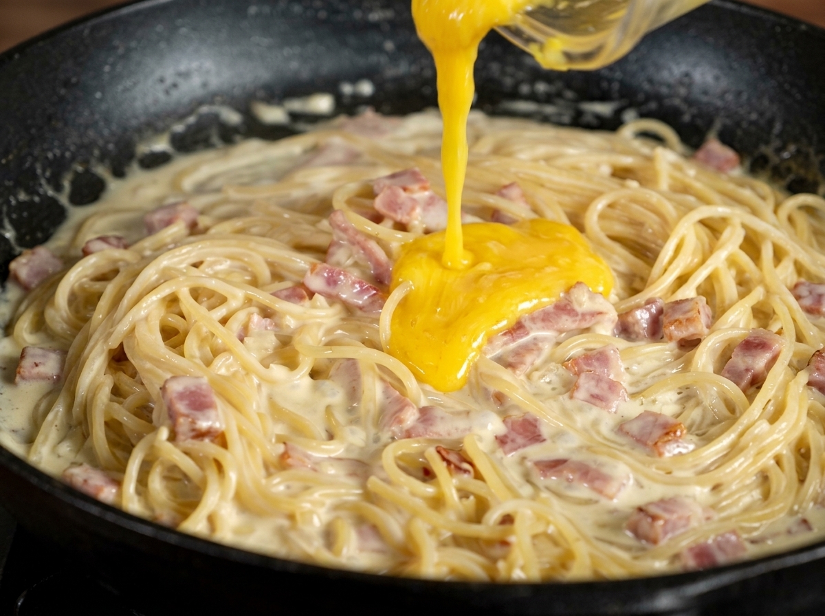 Pasta amarilla de yema y parmesano vertiéndose sobre espaguetis cremosos en una sartén oscura.
