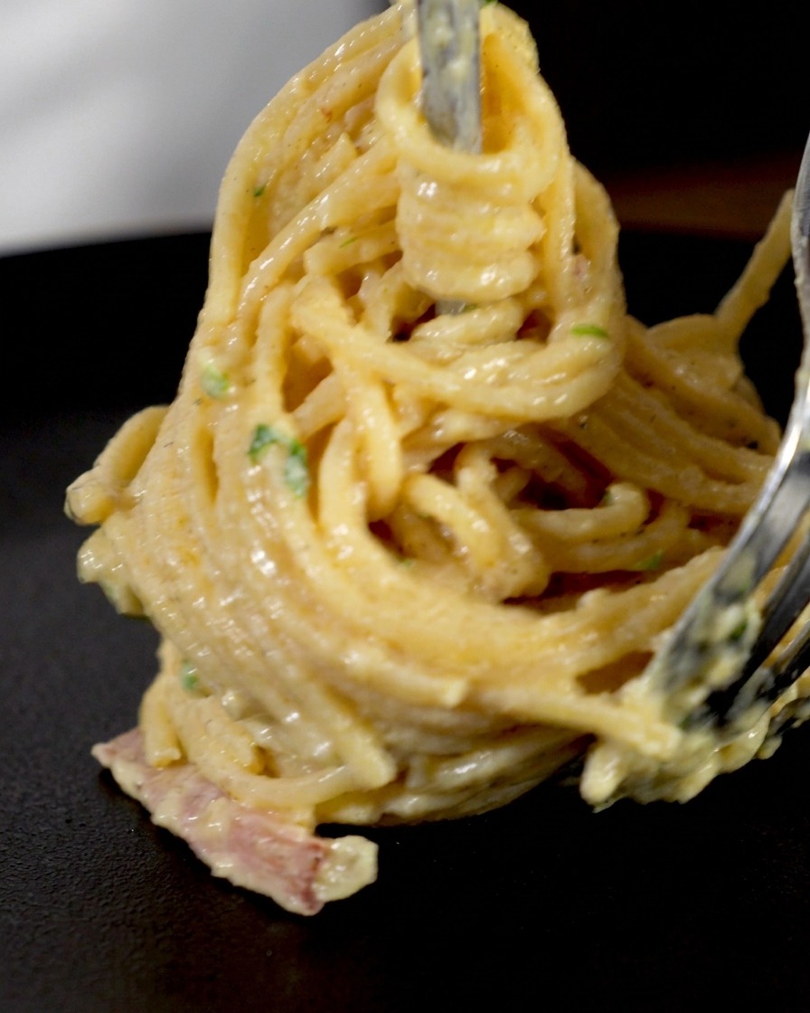 Espaguetis cremosos y trozos de bacon siendo enrollados con un tenedor de trinchar para formar un nido vertical.