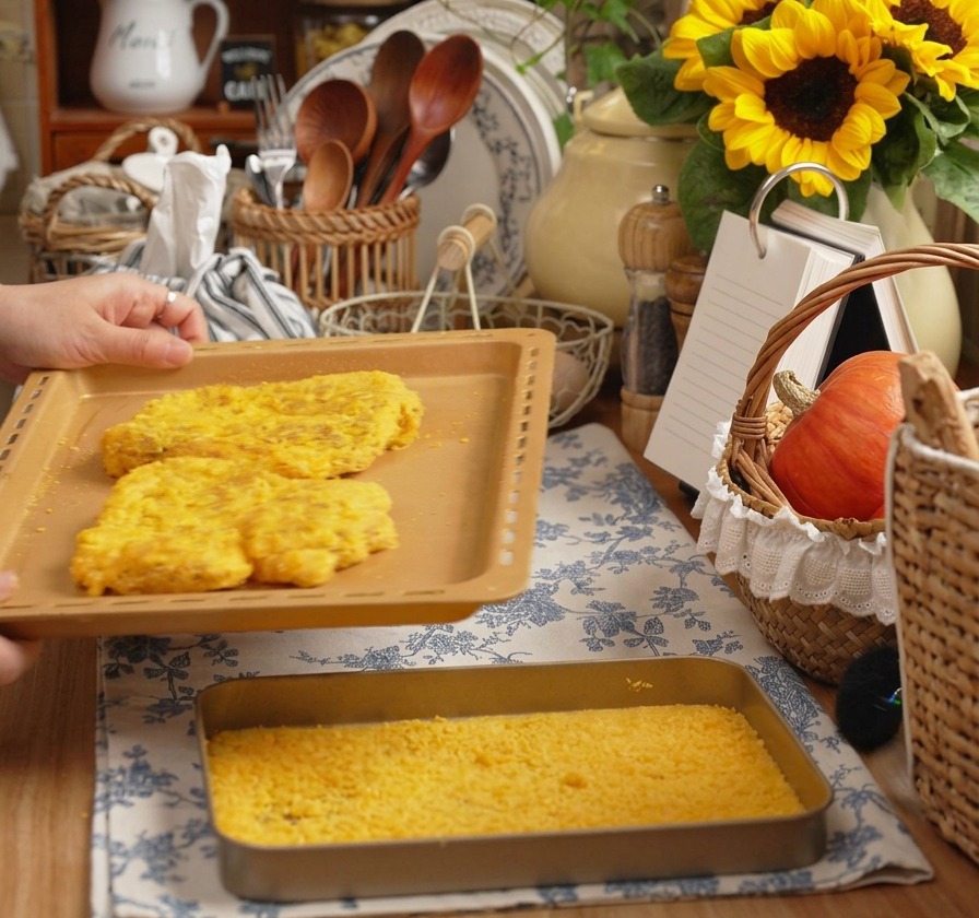 Unas manos sosteniendo una bandeja de horno con dos grandes chuletas de cerdo empanizadas en una cocina acogedora.