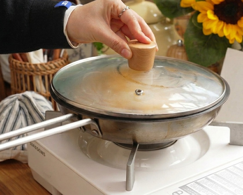 Una mano sosteniendo la perilla de madera de una tapa de vidrio mientras se coloca sobre una sartén de acero inoxidable para cocinar el cerdo y el huevo al vapor.