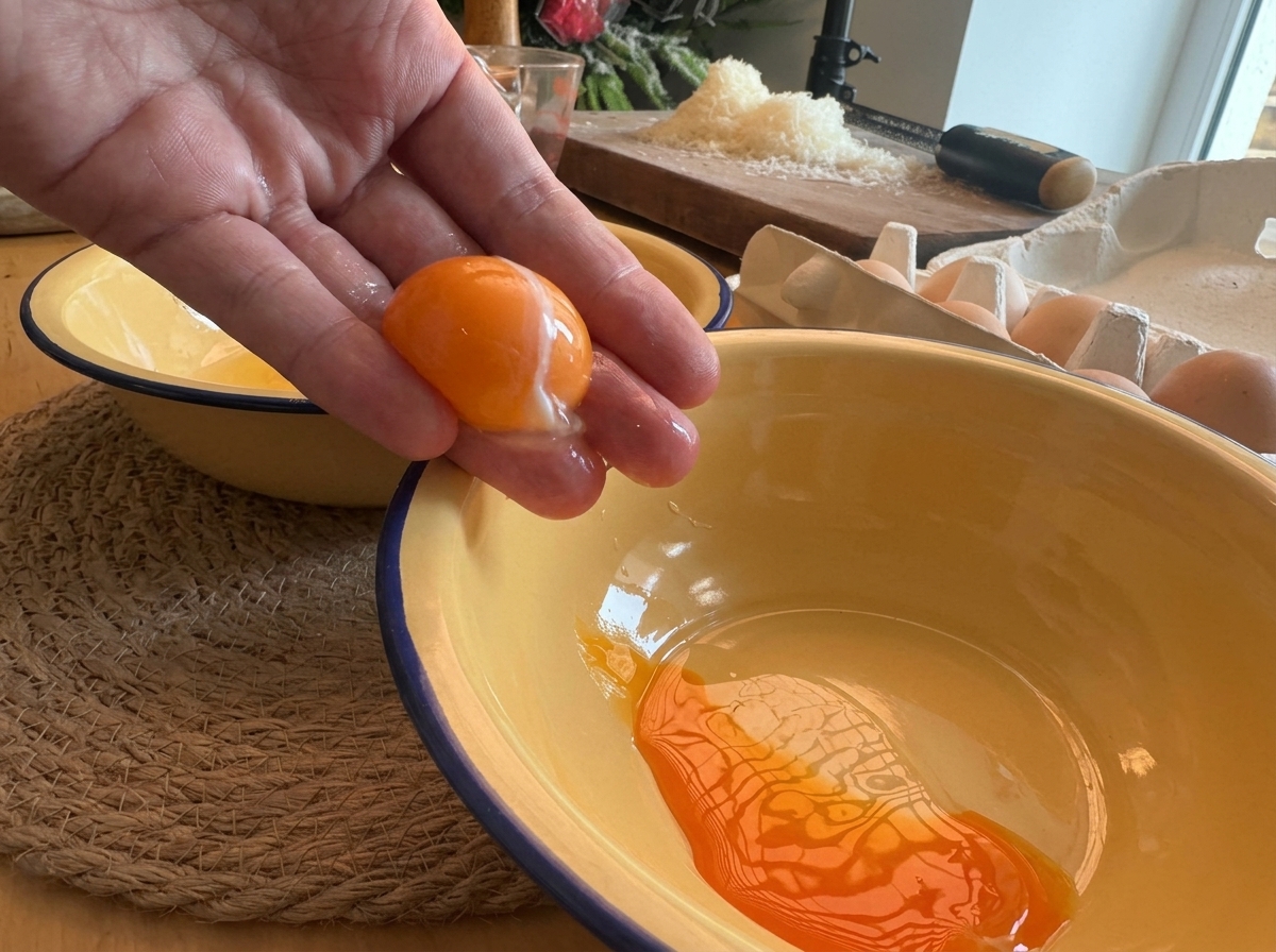 Una mano separando una yema de huevo de color naranja intenso sobre un bol esmaltado amarillo.