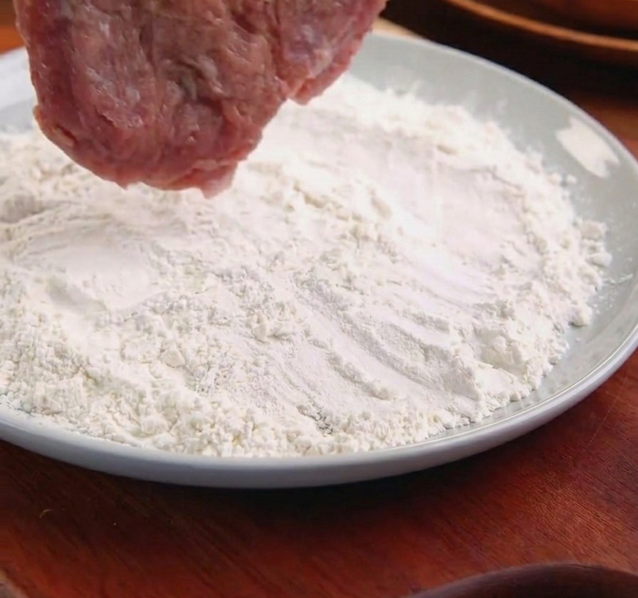 Una chuleta de cerdo sostenida con pinzas de metal siendo colocada en un plato con harina blanca.