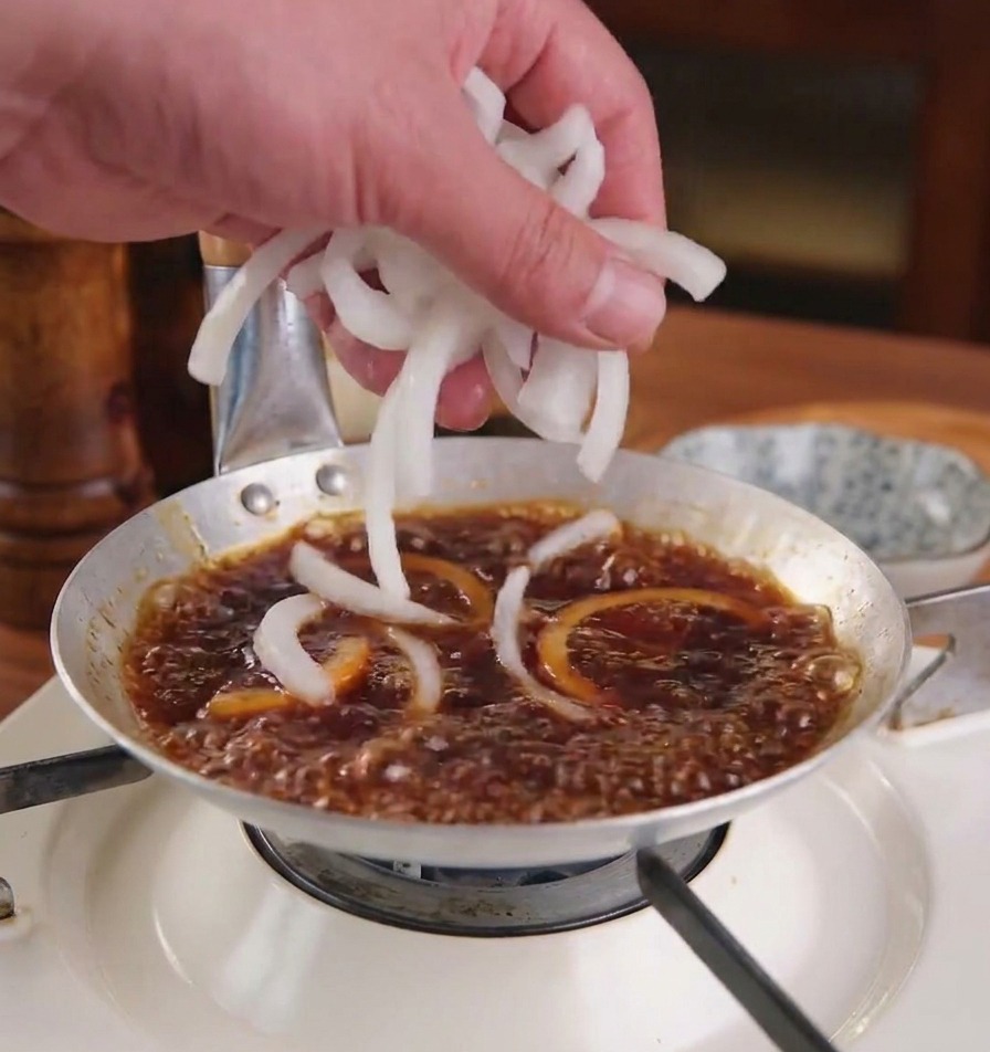 Una mano agregando rodajas de cebolla blanca fresca a una salsa marrón oscuro hirviendo en una sartén metálica baja.