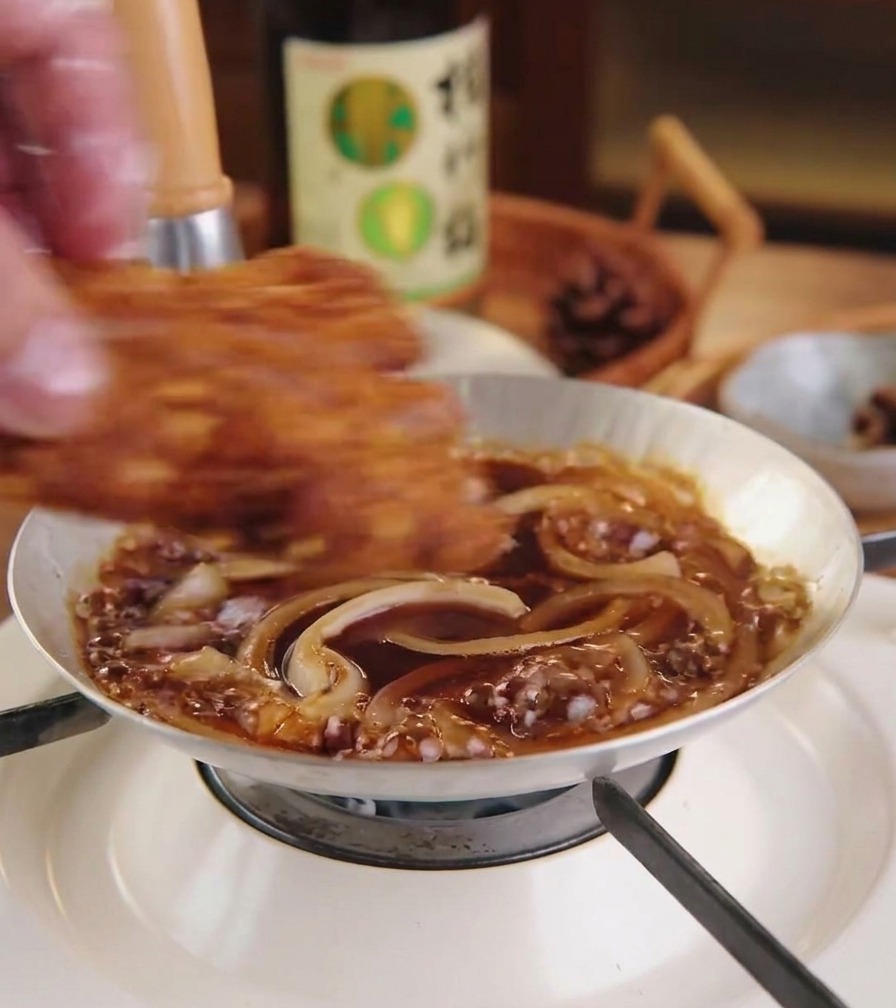 Una chuleta de cerdo dorada y frita colocándose sobre una capa de cebollas y salsa hirviendo en una sartén pequeña.