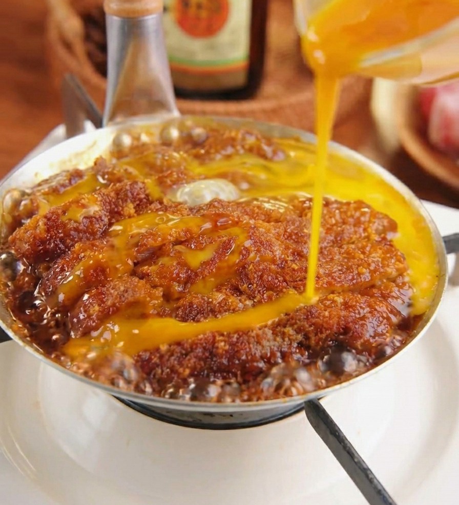 Huevo batido vertiéndose sobre una chuleta de cerdo empanada que se cocina en una salsa oscura con cebollas.