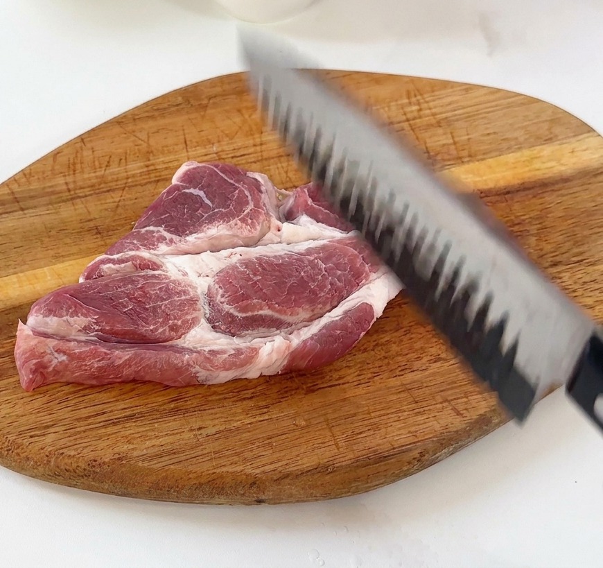 Una mano usando la parte posterior de un cuchillo de chef para golpear y ablandar un filete de cerdo crudo sobre una tabla de madera.