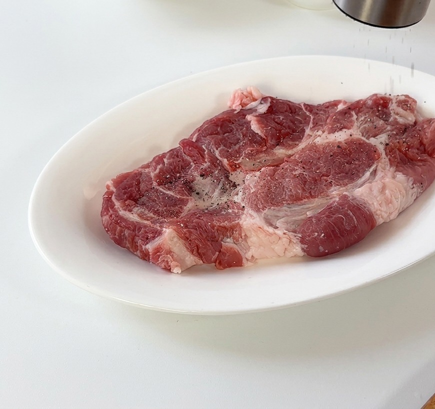 Pimienta negra recién molida cayendo sobre una chuleta de cerdo cruda en un plato de cerámica blanca.
