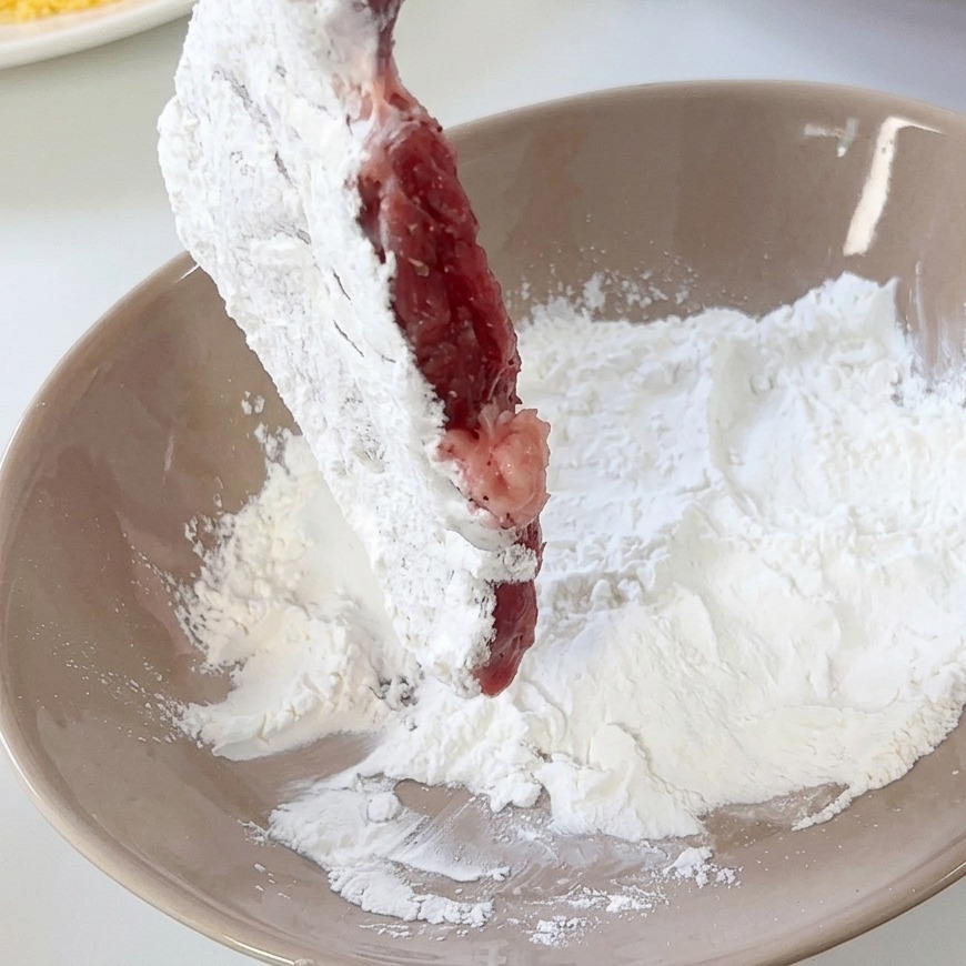 Un filete de cerdo siendo rebozado en harina blanca dentro de un cuenco de cerámica color tostado.