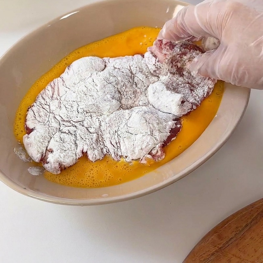 Un filete de cerdo enharinado introduciéndose en un plato hondo con huevo batido amarillo.