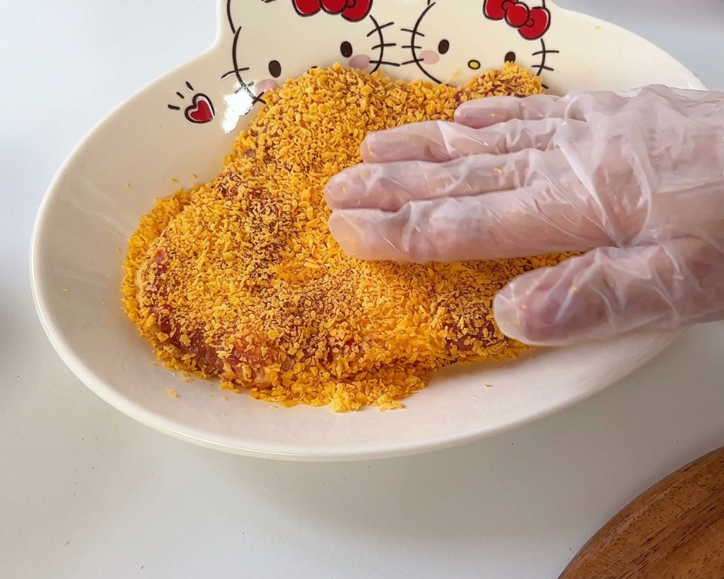 Una mano presionando un filete de cerdo sobre una capa de pan rallado panko en un cuenco con un diseño de Hello Kitty.