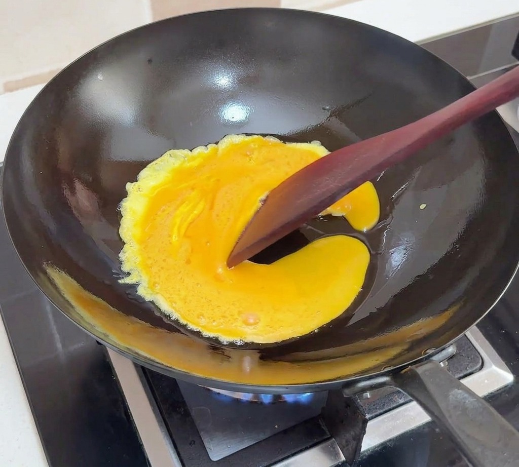 Huevos batidos cocinándose en un wok negro, siendo empujados hacia el centro con una espátula oscura para crear pliegues suaves.