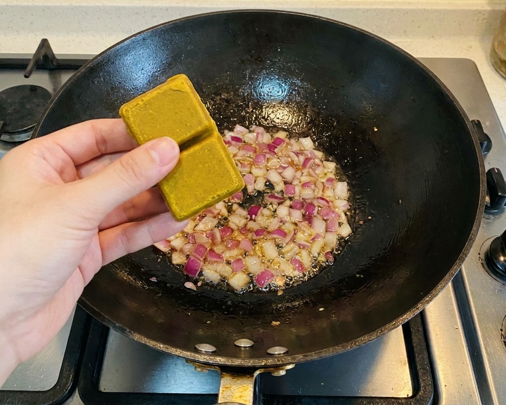 Una persona sosteniendo dos cubos de curry marrón sobre un wok que contiene trozos de cebolla roja salteada.
