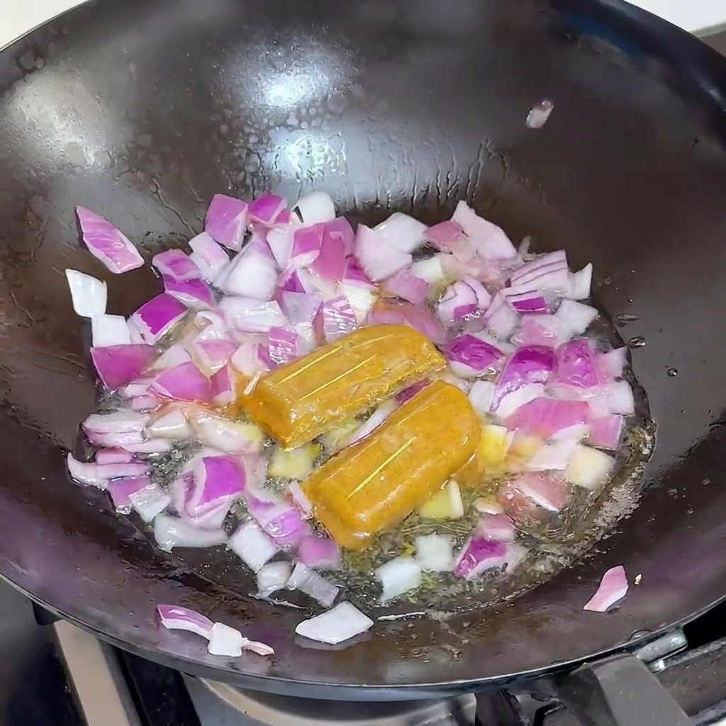 Se está vertiendo agua en un wok negro lleno de cebolla roja y dos cubos de curry enteros.