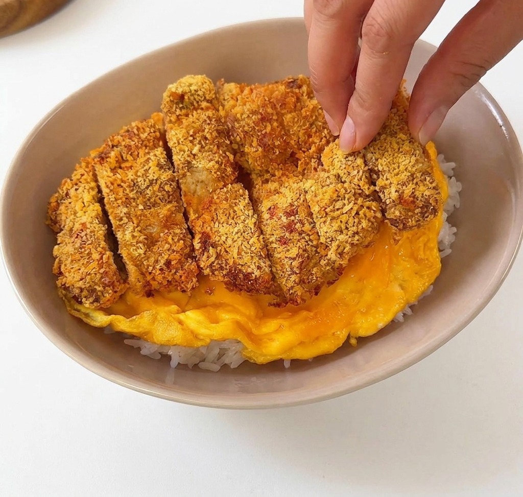 Chuleta de cerdo empanada y cortada siendo colocada encima de una tortilla amarilla suave dentro de un cuenco de cerámica beige con arroz blanco.