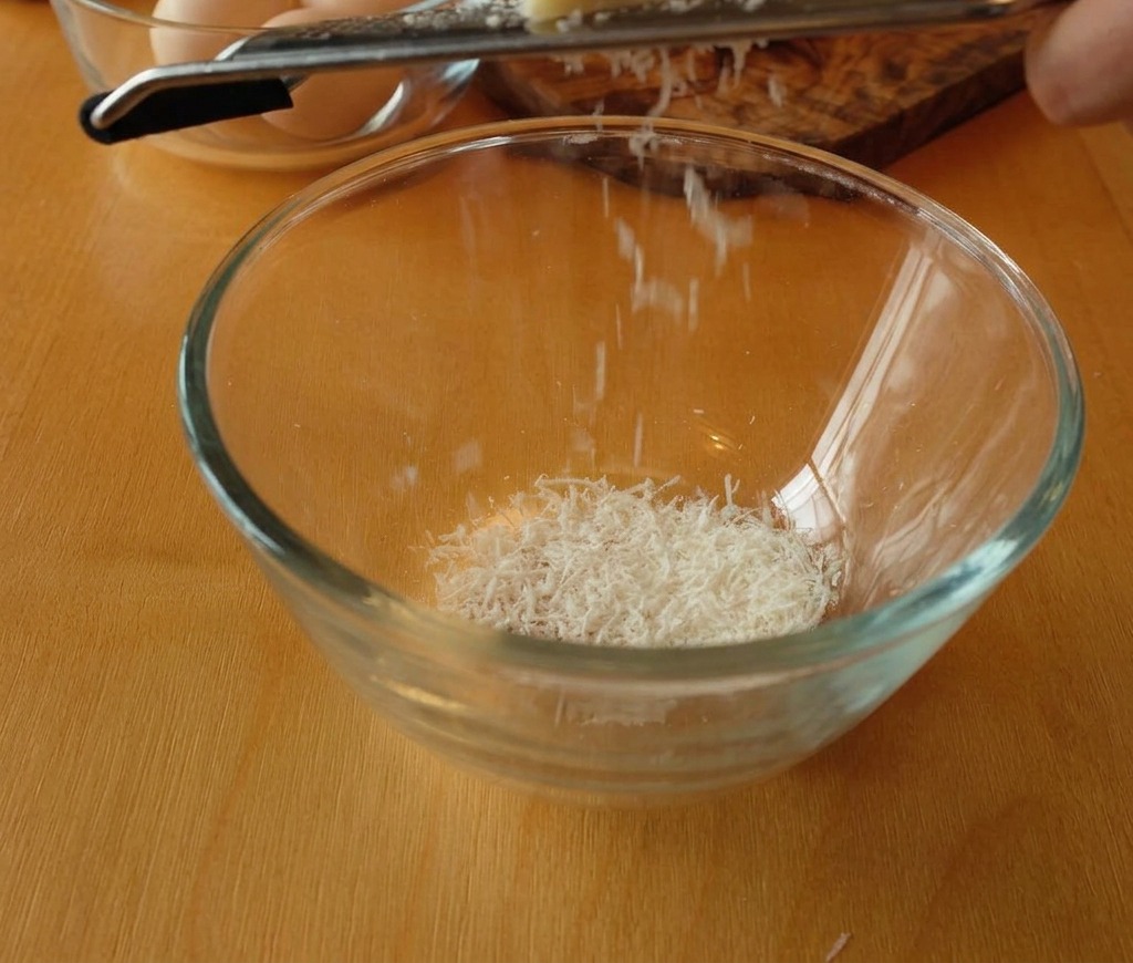 Un bloque de queso Pecorino Romano siendo rallado en un bol de cristal, creando una montaña de virutas blancas finas.