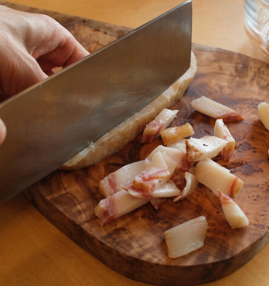 Un cuchillo afilado retirando la piel gruesa y oscura de un trozo de guanciale sobre una tabla de madera.