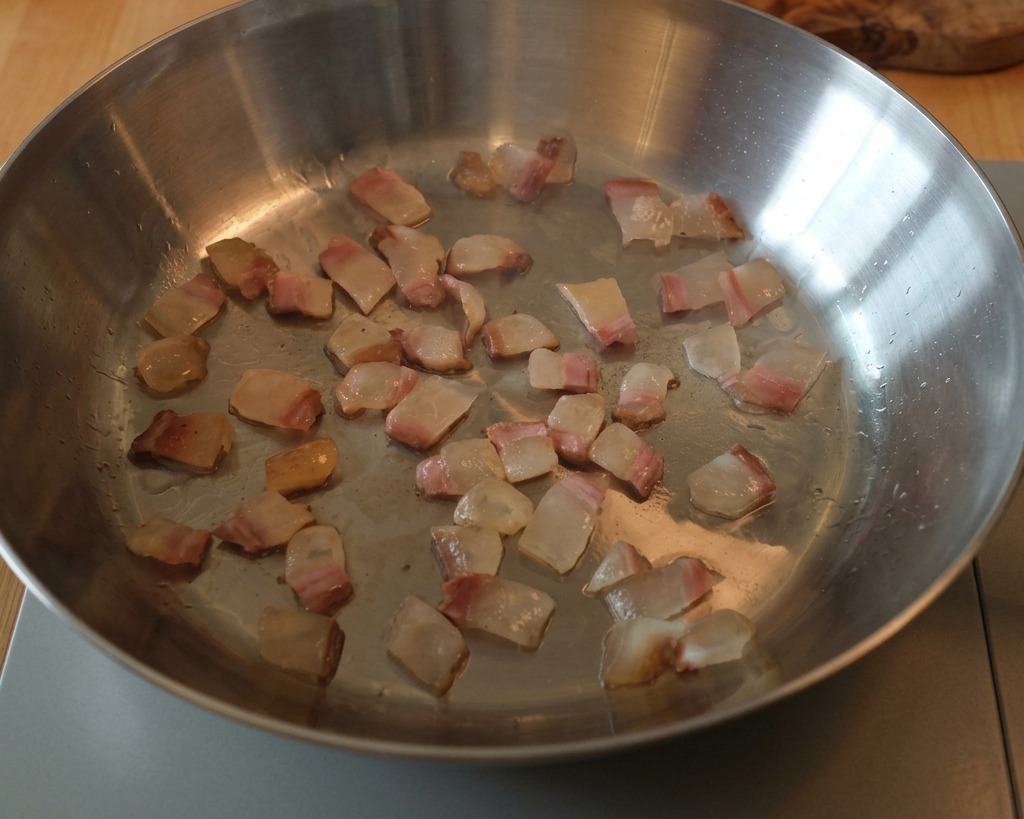 Dados de guanciale dorándose en una sartén de acero, con la grasa convirtiéndose en un líquido transparente.