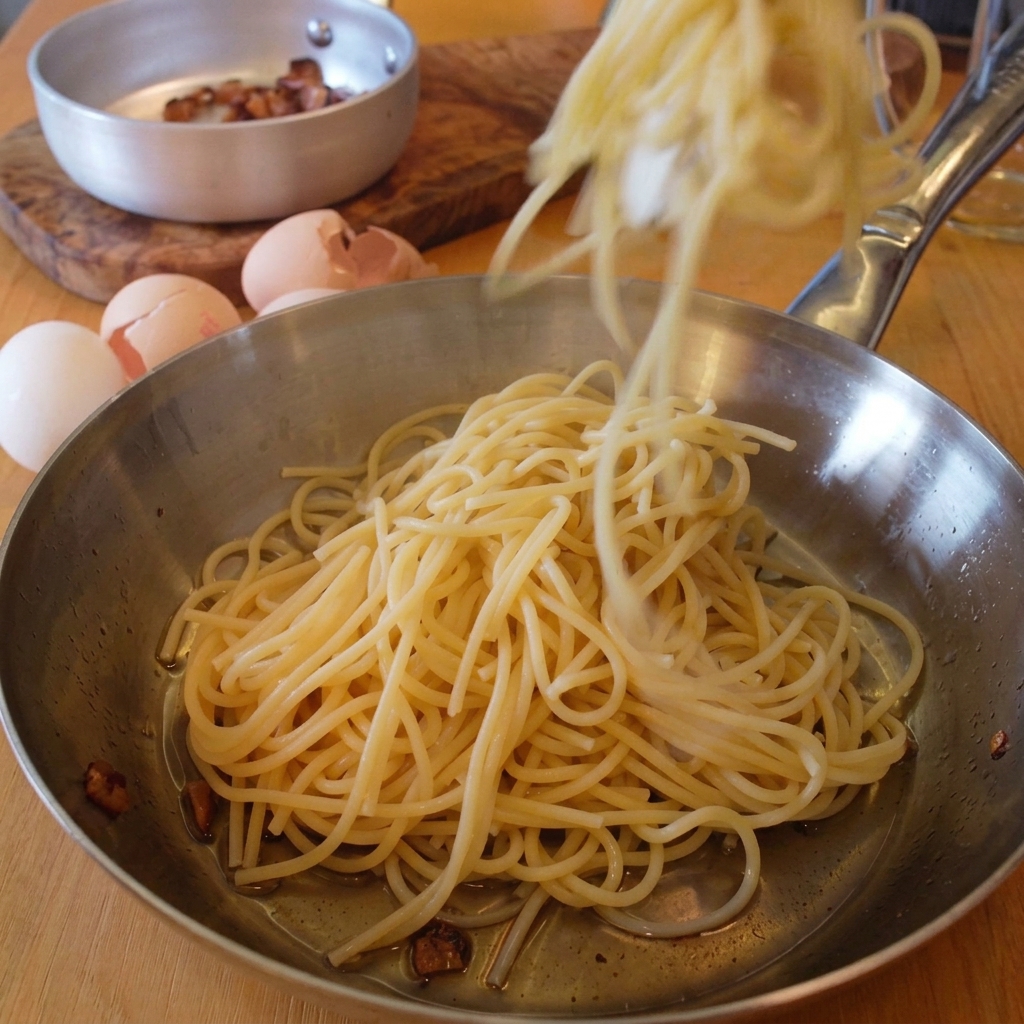 Pinzas dejando caer espaguetis recién cocidos en una sartén de acero con restos de grasa de guanciale.