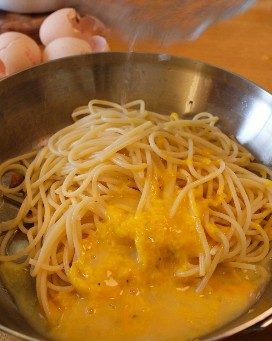 Espaguetis en una sartén cubiertos de salsa amarilla mientras se vierte un chorrito de agua de la pasta por encima.