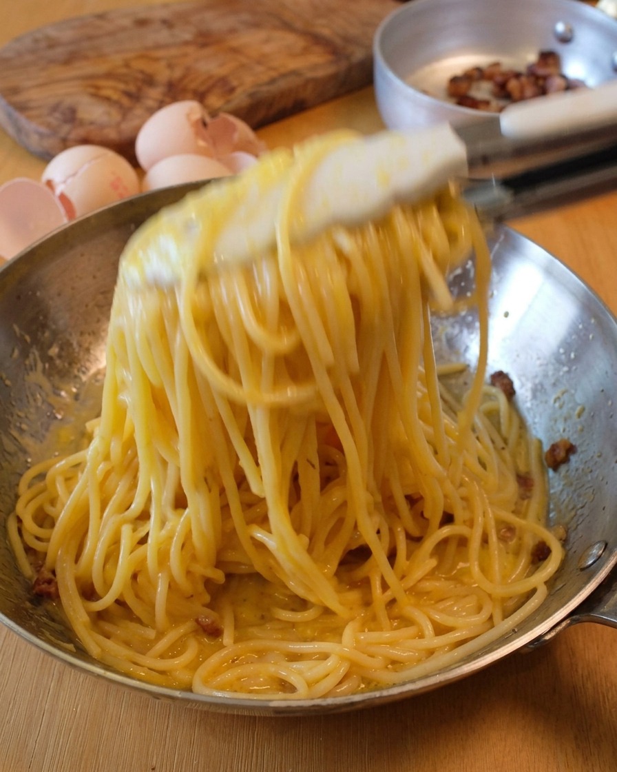 Salteando espaguetis en una sartén metálica con pinzas para crear la emulsión de la salsa carbonara.