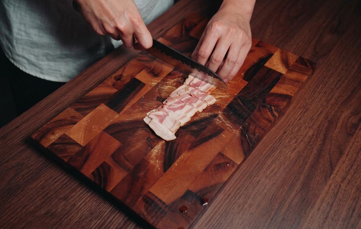 Una persona cortando tiras de beicon crudo en pequeños rectángulos sobre una tabla de madera oscura.