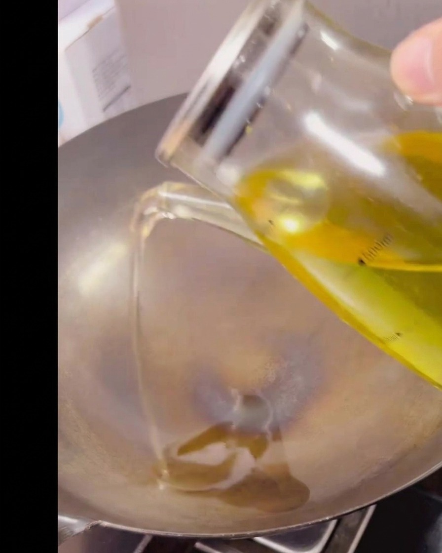 Aceite de cocina transparente vertiéndose desde una jarra medidora de cristal en un wok de metal caliente y vacío.