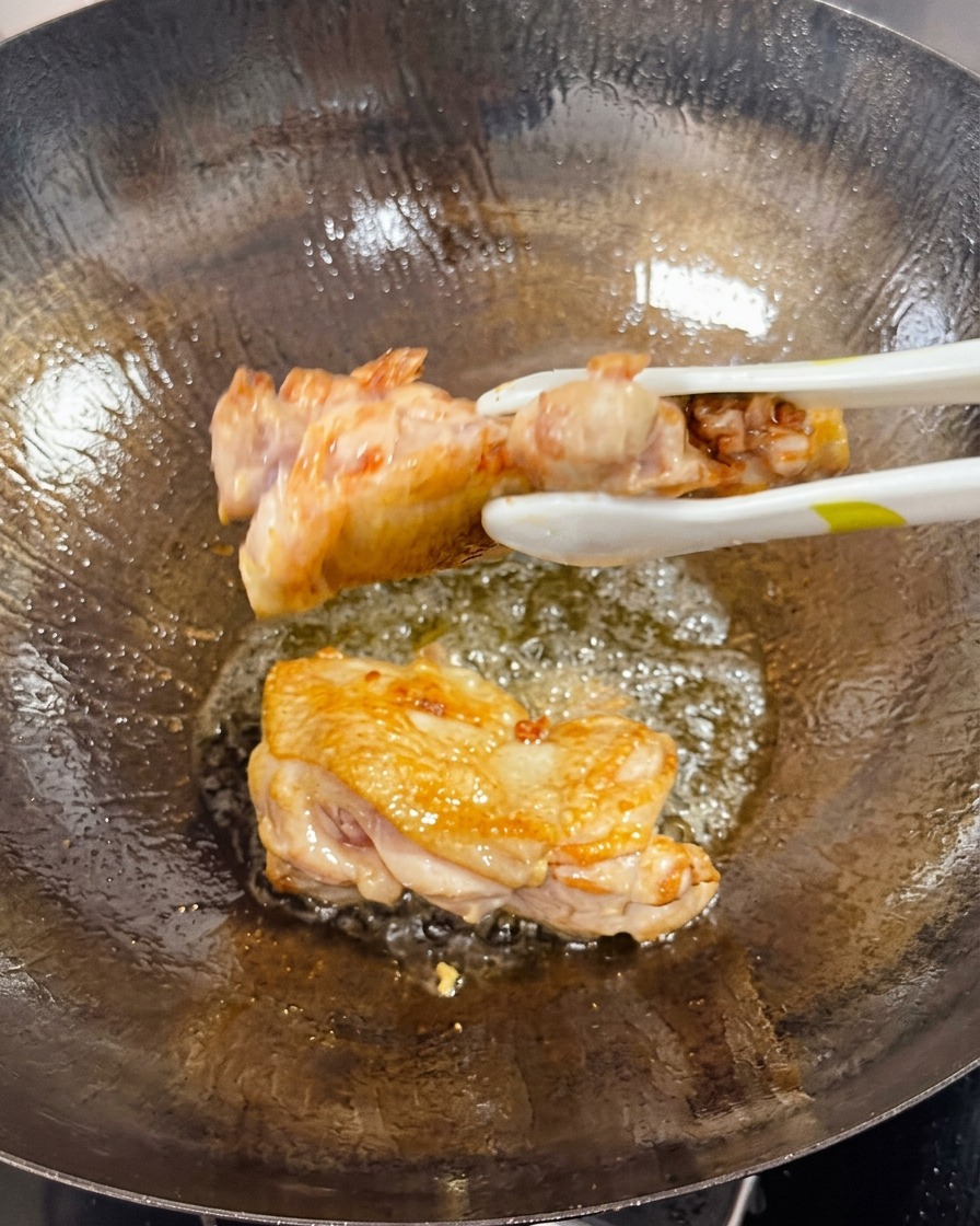 Pinzas dando la vuelta a un muslo de pollo sellado en un wok, mostrando el lado cocinado dorado y crujiente.