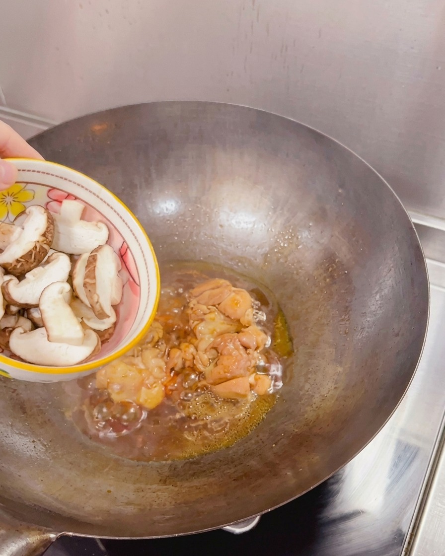 Setas shiitake frescas en rodajas añadiéndose desde un bol a un wok que contiene pollo sellado y salsa hirviendo a fuego lento.