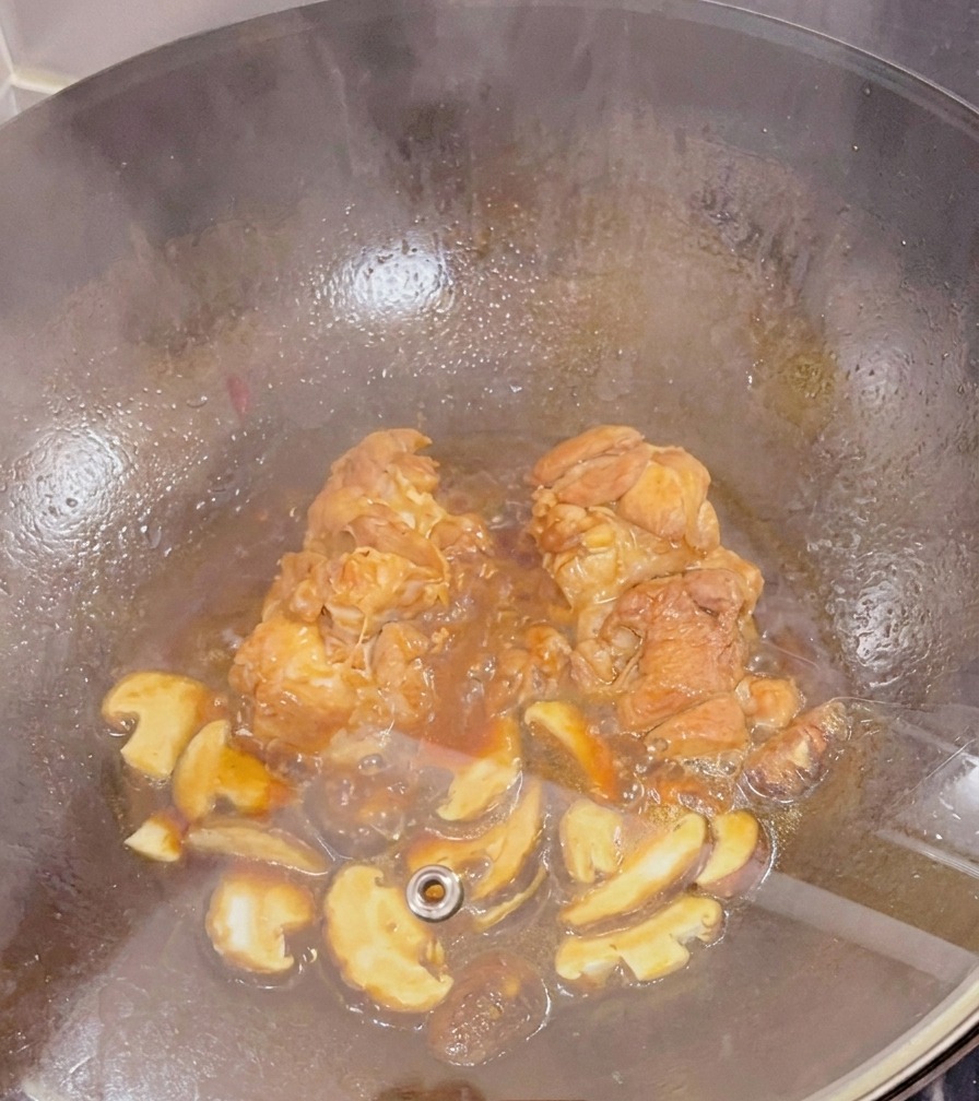 Muslos de pollo y setas shiitake en rodajas cocinándose a fuego lento en una salsa oscura dentro de un wok bajo una tapa de cristal.