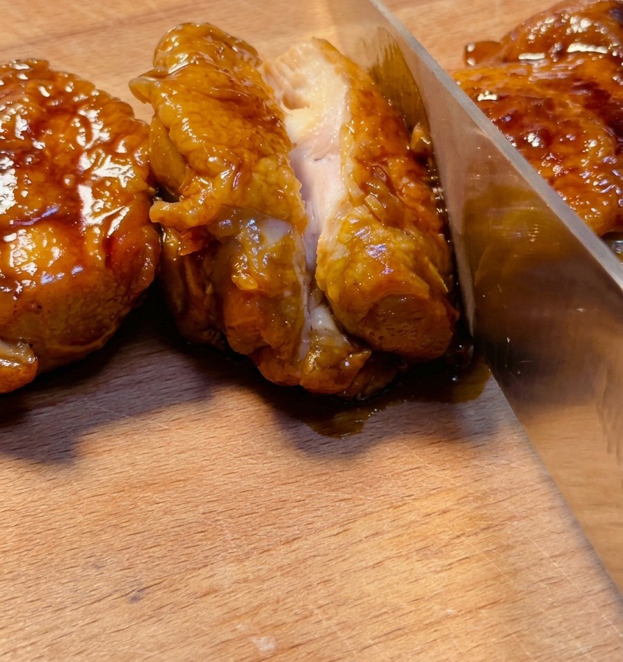 Un cuchillo afilado cortando a través de un muslo de pollo teriyaki profundamente glaseado en una tabla de madera, mostrando la jugosa carne en su interior.