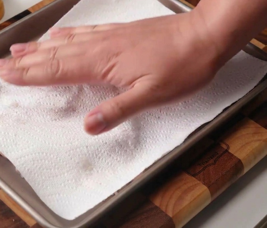 Una mano usando una toalla de papel blanca para secar contramuslos de pollo crudos colocados en una bandeja para hornear de metal.