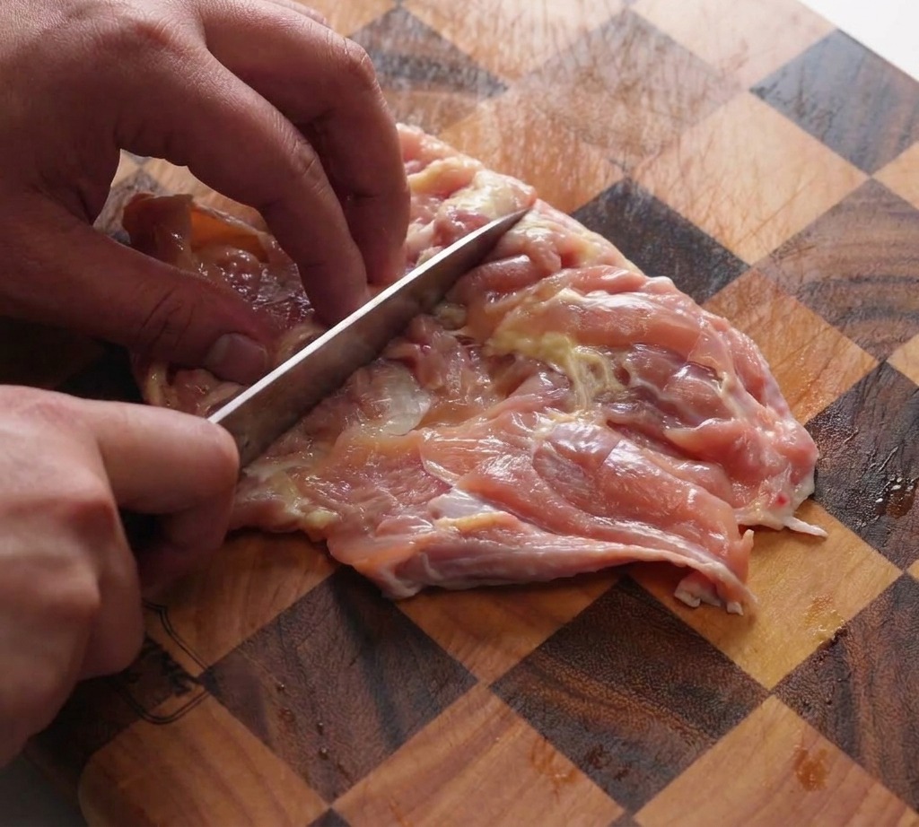 Una mano utilizando el lomo del cuchillo de un chef para golpear contramuslos de pollo deshuesados sobre una tabla de cortar de madera a cuadros.