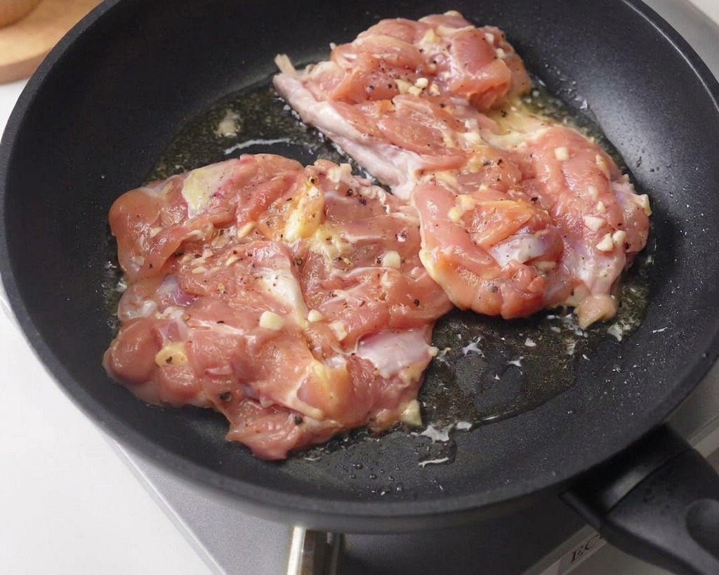 Dos contramuslos de pollo marinados dorándose con la piel hacia abajo en una sartén antiadherente oscura.