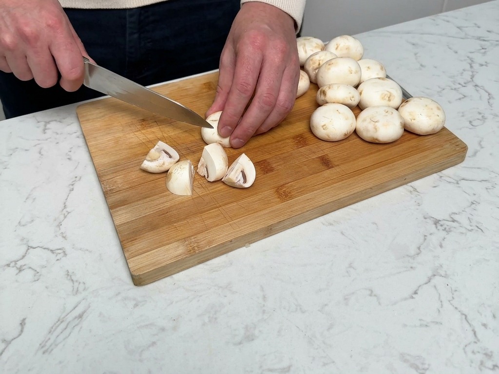 Manos usando un cuchillo de chef para cortar champiñones blancos en cuartos sobre una tabla de madera.