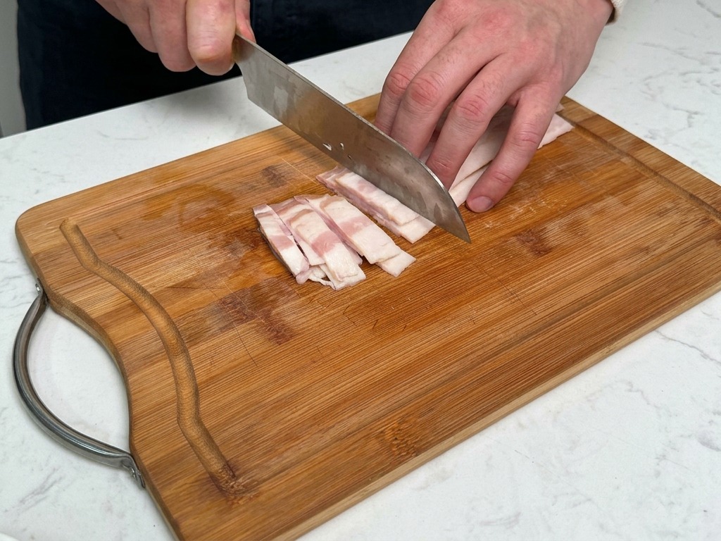 Una persona cortando tiras gruesas de beicon en una tabla de madera con un cuchillo.