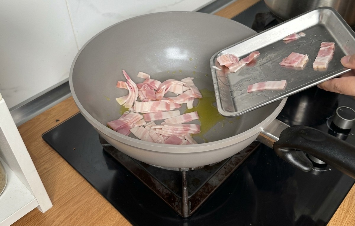 Deslizando tiras de beicon crudo desde una bandeja metálica a una sartén gris con aceite de oliva.