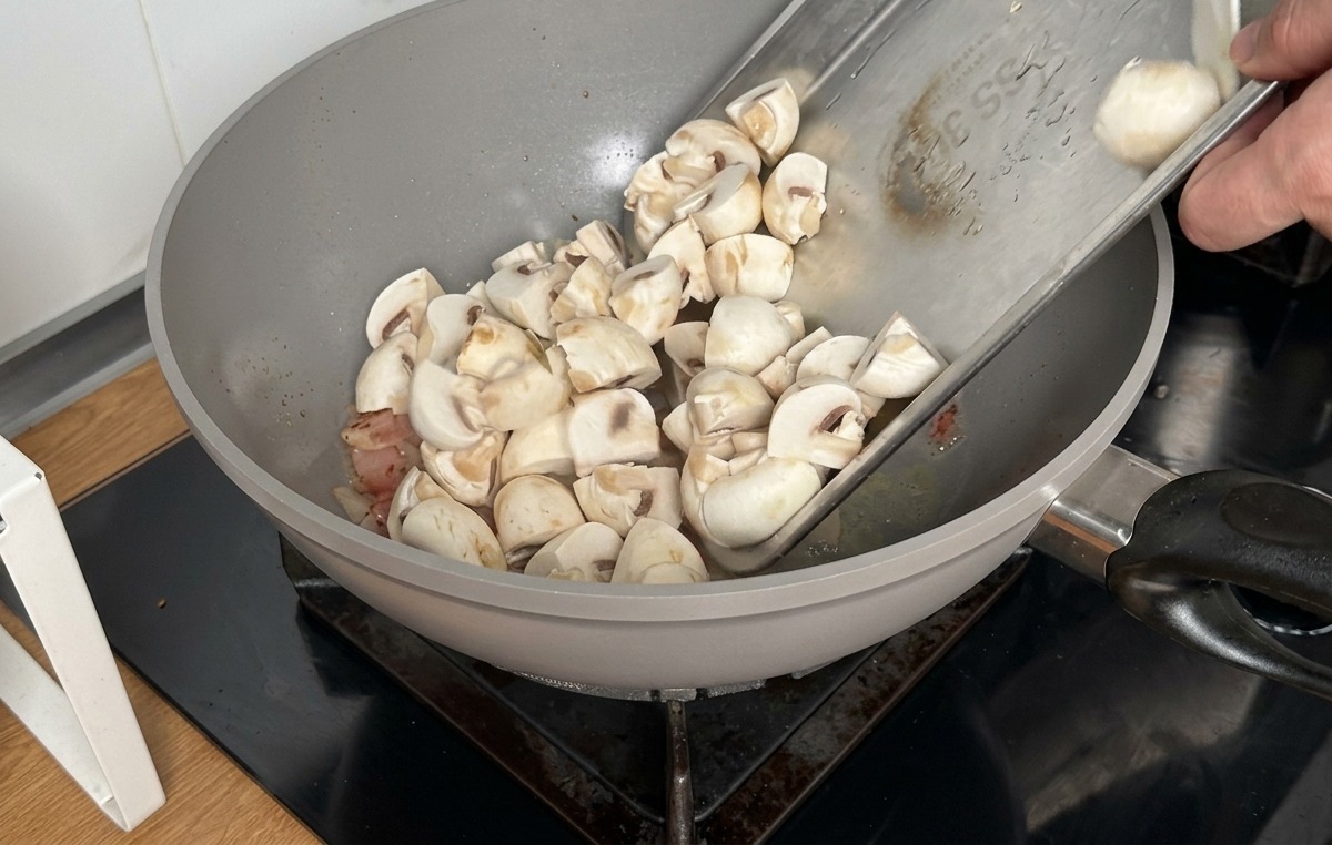 Vertiendo champiñones blancos troceados desde una bandeja metálica a un wok con el beicon cocinado.